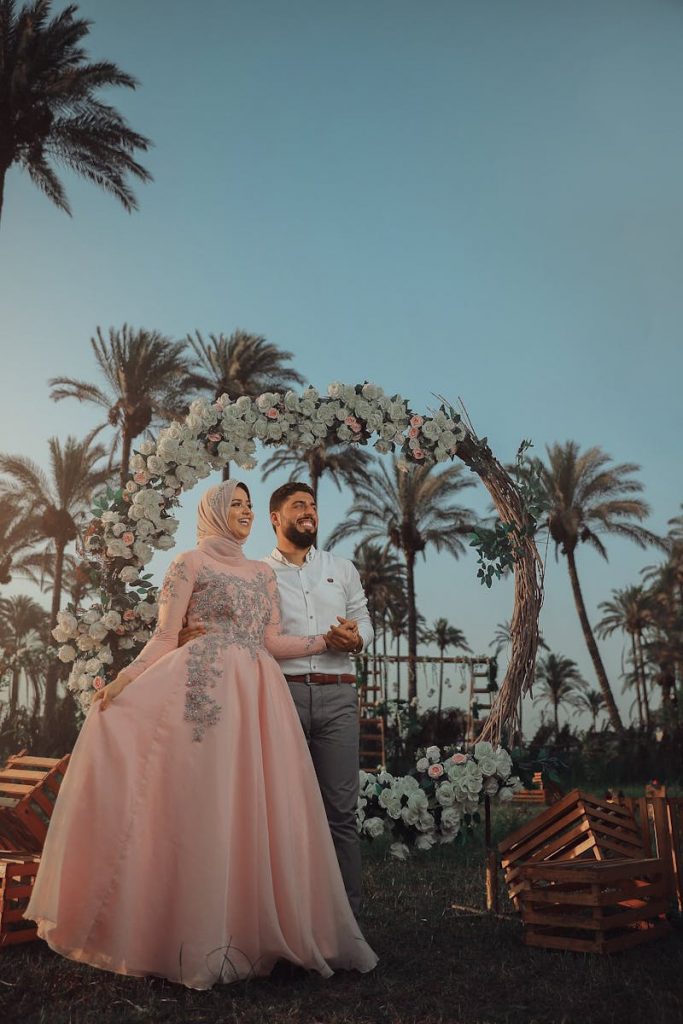 pexels-photo-16800881-16800881 Bride and groom in traditional attire under floral arch with palm trees.