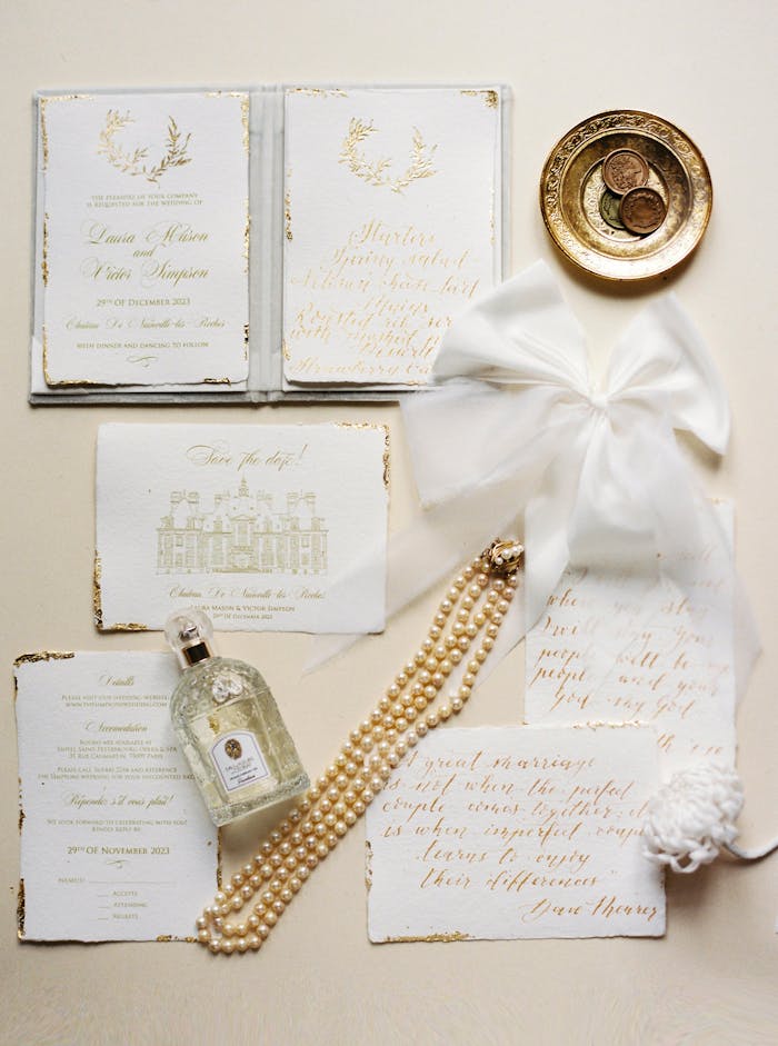 gallery-2 Sophisticated wedding stationery flat lay with perfume, pearls, coins, and a white ribbon on a neutral background.