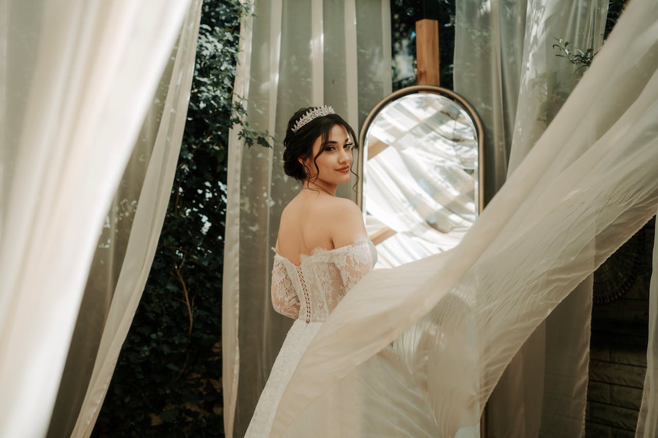 gallery-1 A beautiful bride in a lace wedding dress looks back towards a mirror surrounded by flowing curtains.